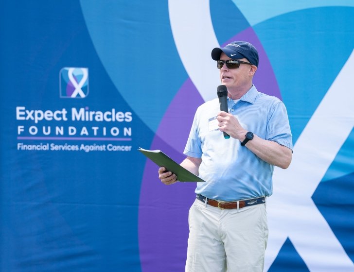 Man speaking into a microphone in front of an Expect Miracles Foundation backdrop
