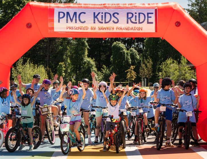 Group of young bike riders stand with arms in the air under 'PMC Kids Ride' start line
