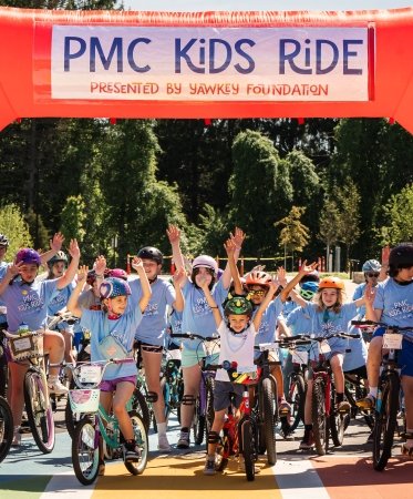 Group of young bike riders stand with arms in the air under “PMC Kids Ride” start line