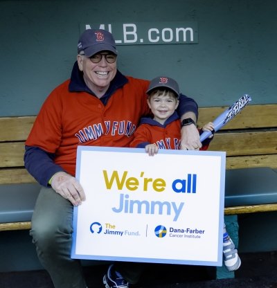 Granddad and grandson in Fenway dugout with We’re All Jimmy sign