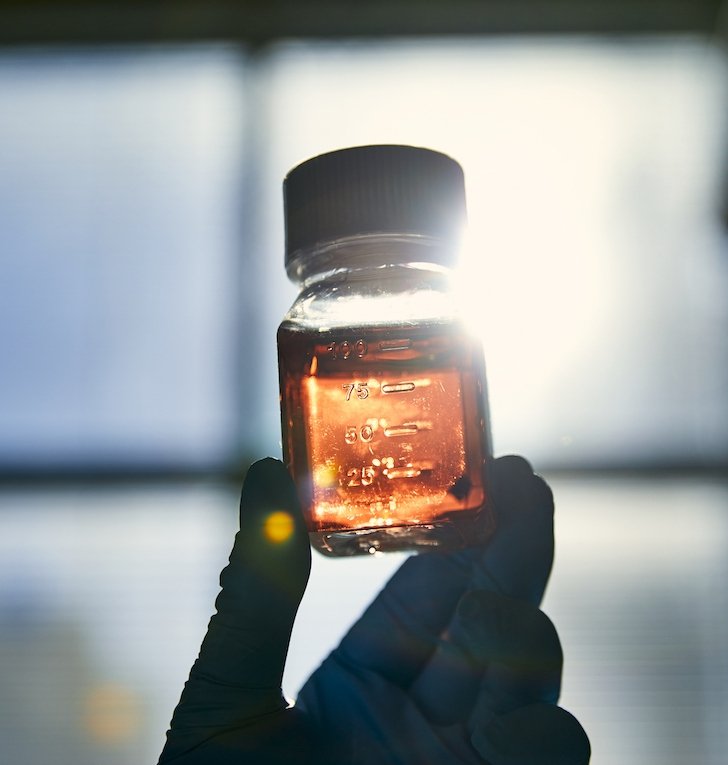 A gloved hand holds up a bottle full of liquid in front of the sunny window of a lab room