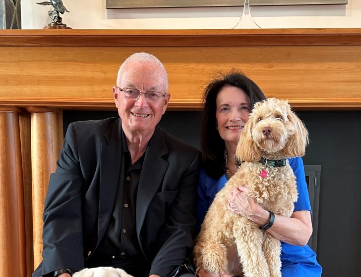 Larry and Pam sit in chairs in front of a fireplace mantel, holding 2 dogs