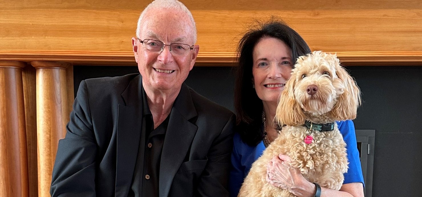 Larry and Pam sit in chairs in front of a fireplace mantel, holding 2 dogs
