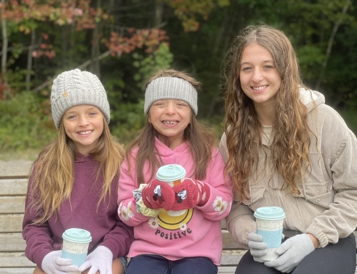Three sisters sit on bench holding drinks with trees in background