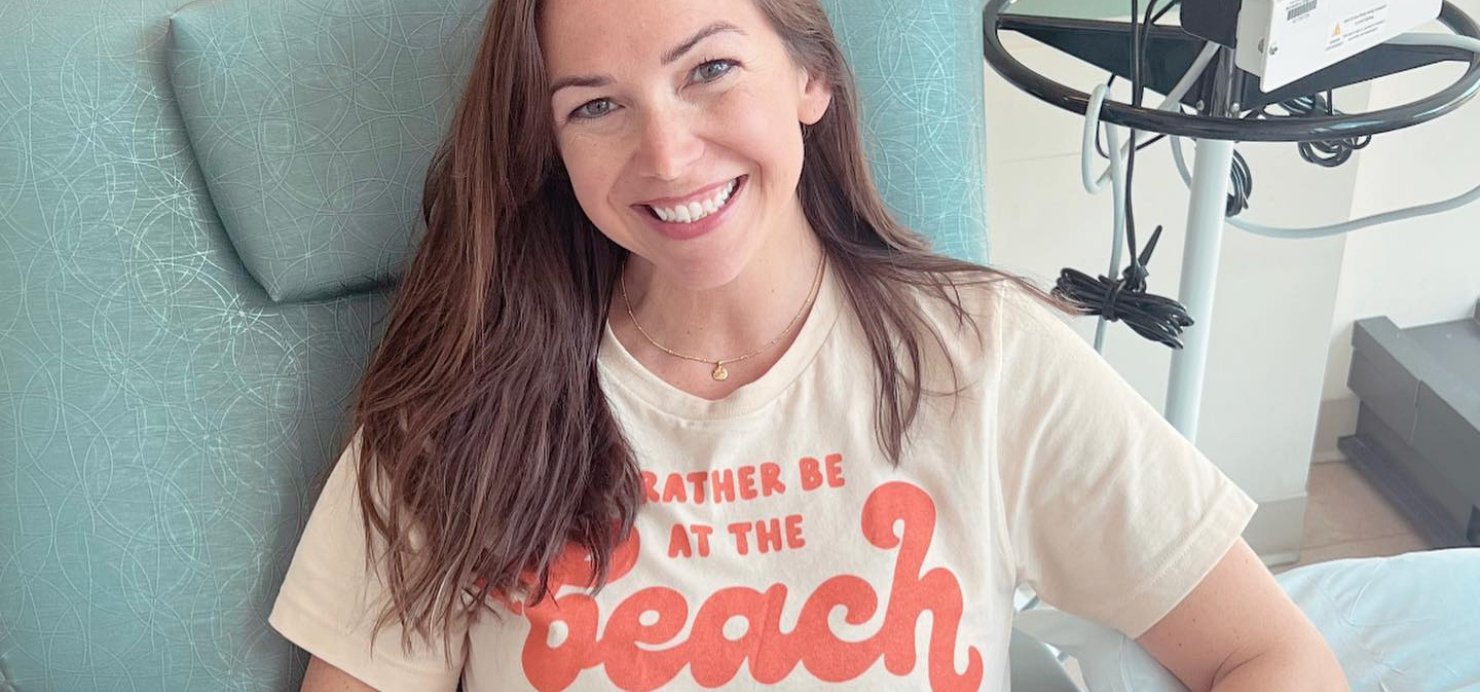 Julie sits in a hospital bed smiling wearing 'I’d rather be at the beach' T-shirt