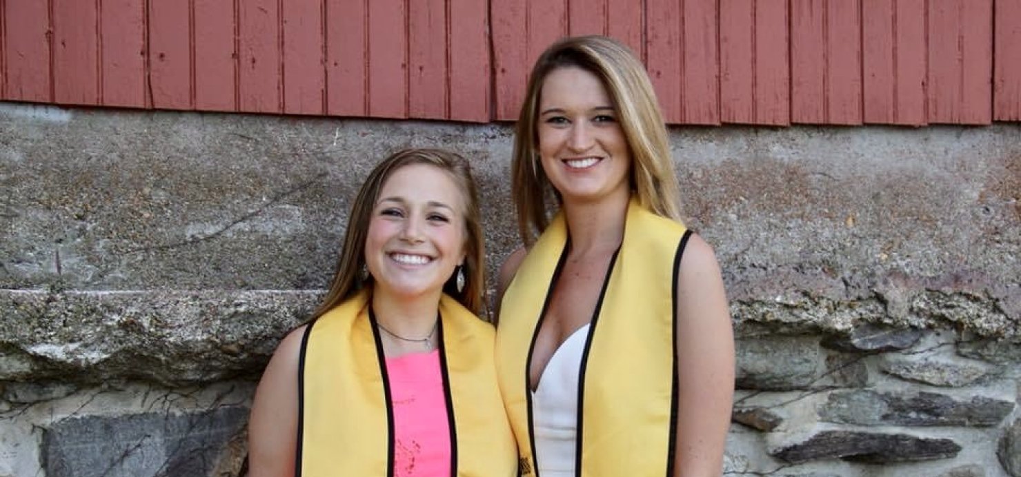 Grace and Gabby stand together in front of a wall wearing graduation clothes