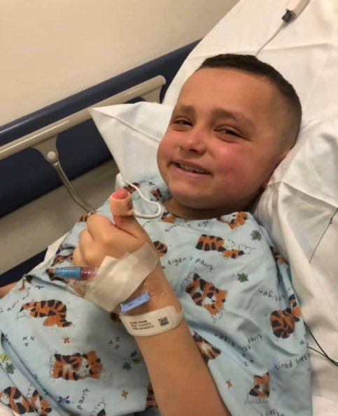 Grady, wearing a hospital gown in a hospital bed, giving a thumbs up