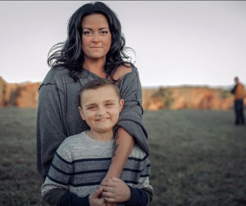 Mother stands behind son in a field with a background of trees