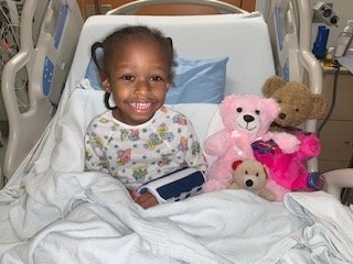 Samara sits in a hospital bed surrounded by stuffed animals