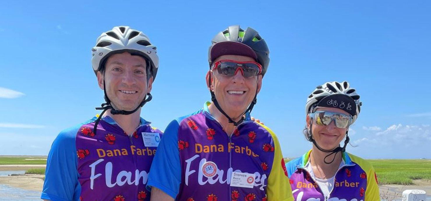 Three bike riders pose in front of a marsh while wearing helmets and 'Dana-Farber Flames' jerseys