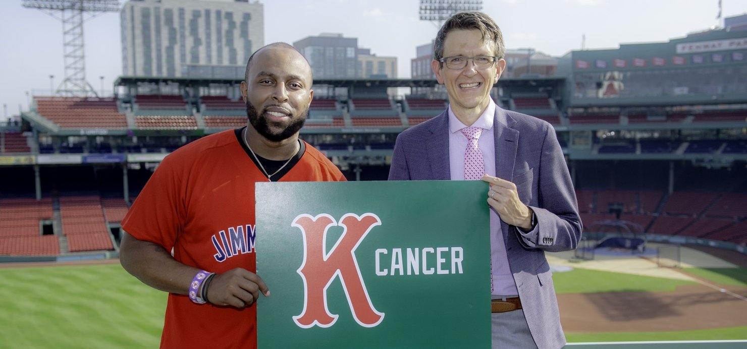 Lamar stands with a doctor in front of Fenway Park’s Green Monster, holding a K Cancer sign