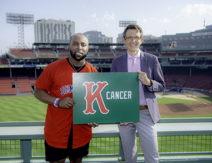Lamar stands with a doctor in front of Fenway Park’s Green Monster, holding a K Cancer sign