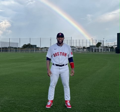 Lamar stands on a baseball field wearing Red Sox jersey with a rainbow in the sky behind him