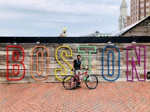 Benson stands in front of rainbow 'Boston' sign, holding his bike