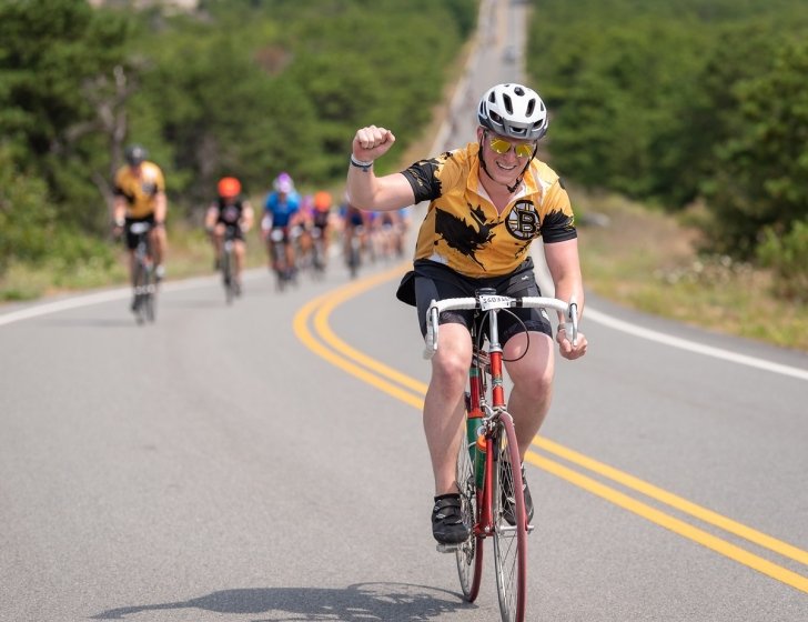 Sam rides a bike on a road with his fist in the air and other bikers behind him