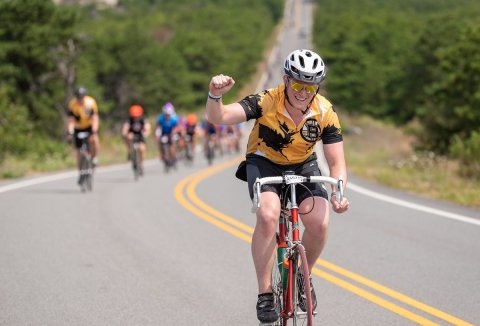 Sam rides bike on a road with his fist in the air and other bikers behind him