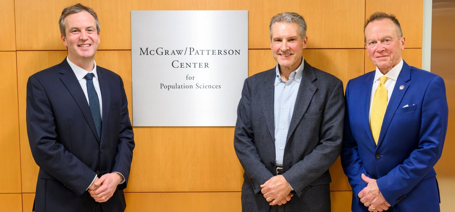 McGraw and Patterson families stand next to 'McGraw/Patterson Center for Population Sciences' wall sign