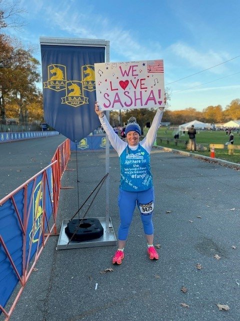 Beth stands at the B.A.A. Half Marathon holding up a sign reading 'We love Sasha'